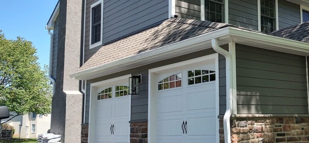 White seamless gutter and downspout along a garage roof, with gray lap siding and stone
